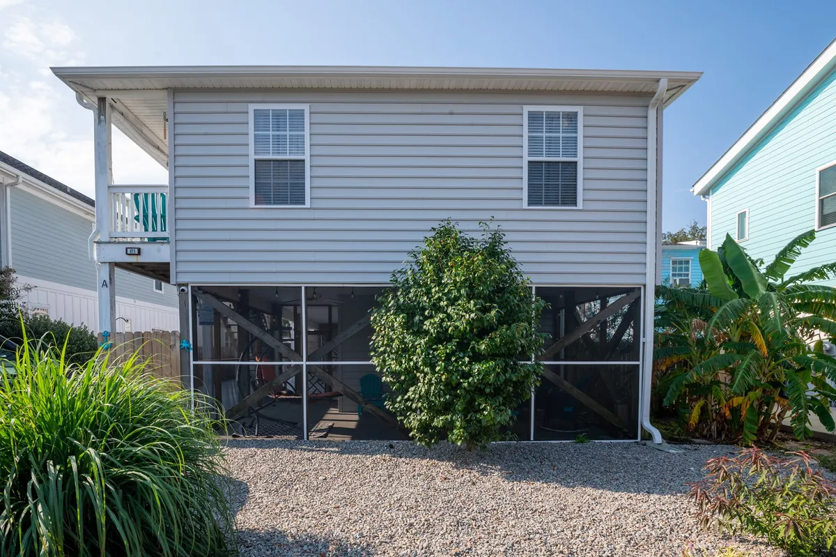 An inviting exterior view of the duplex, showcasing its elevated structure surrounded by lush greenery and a gravel pathway. The spacious porch hints at spots for group gatherings and relaxation outdoors, while the tranquil garden setting suggests a peaceful retreat ideal for families.