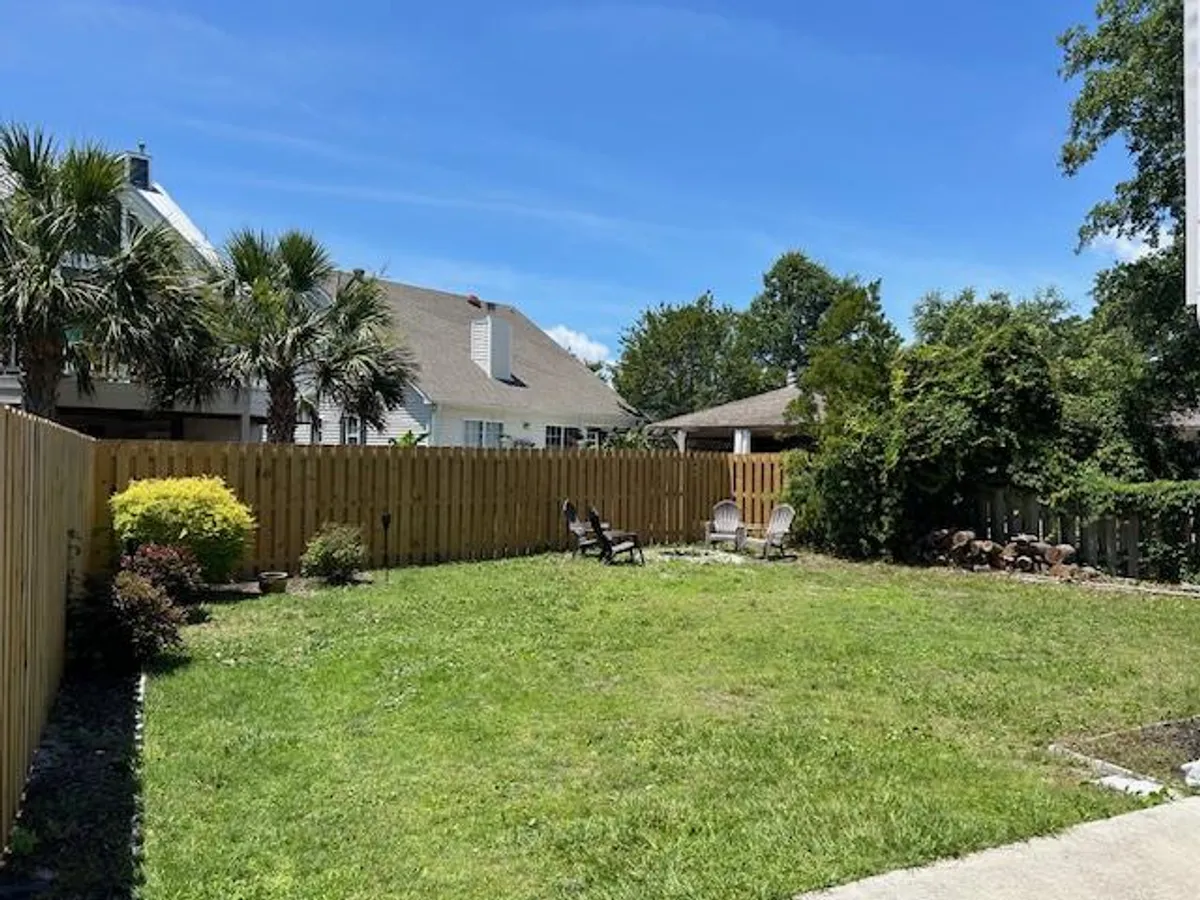 A vibrant backyard scene with green grass, palm trees, wooden fence, and lawn chairs set up for outdoor relaxation. The space encourages group activities like backyard games, dining al fresco, or morning coffee together, encapsulating the spirit of multi-generational gatherings.