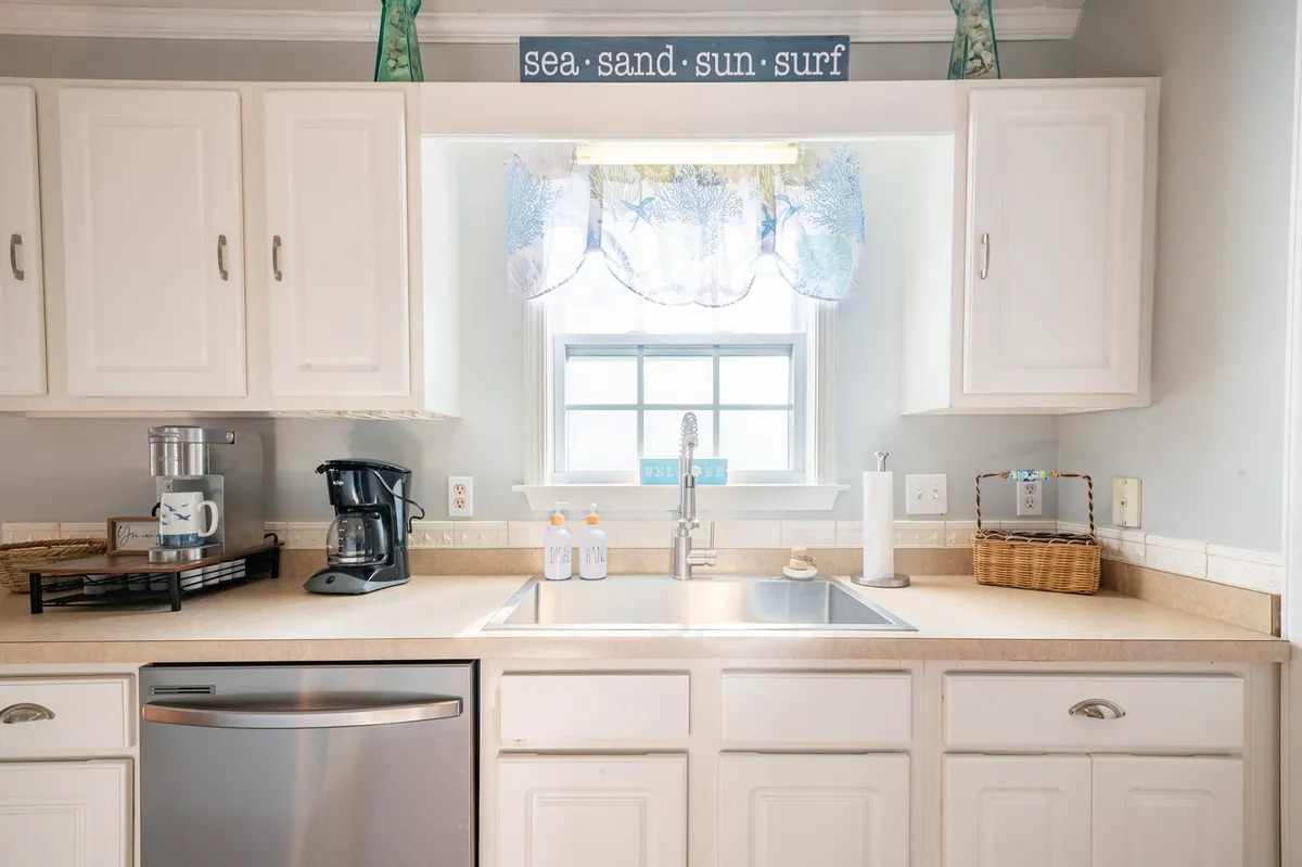 A cheerful kitchen scene featuring a kitchen sink, coffee maker, and a window with sea-themed curtains. A decorative sign and inviting countertop space evoke a sense of morning rituals and easy meal prep before setting out for a day of exploration.