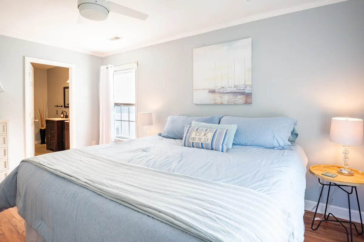 A spacious primary bedroom with a large bed dressed in blue bedding, complemented by a painting of boats above the headboard and nightstands with lamps on either side. The room is softly lit, evoking a tranquil, restful atmosphere.