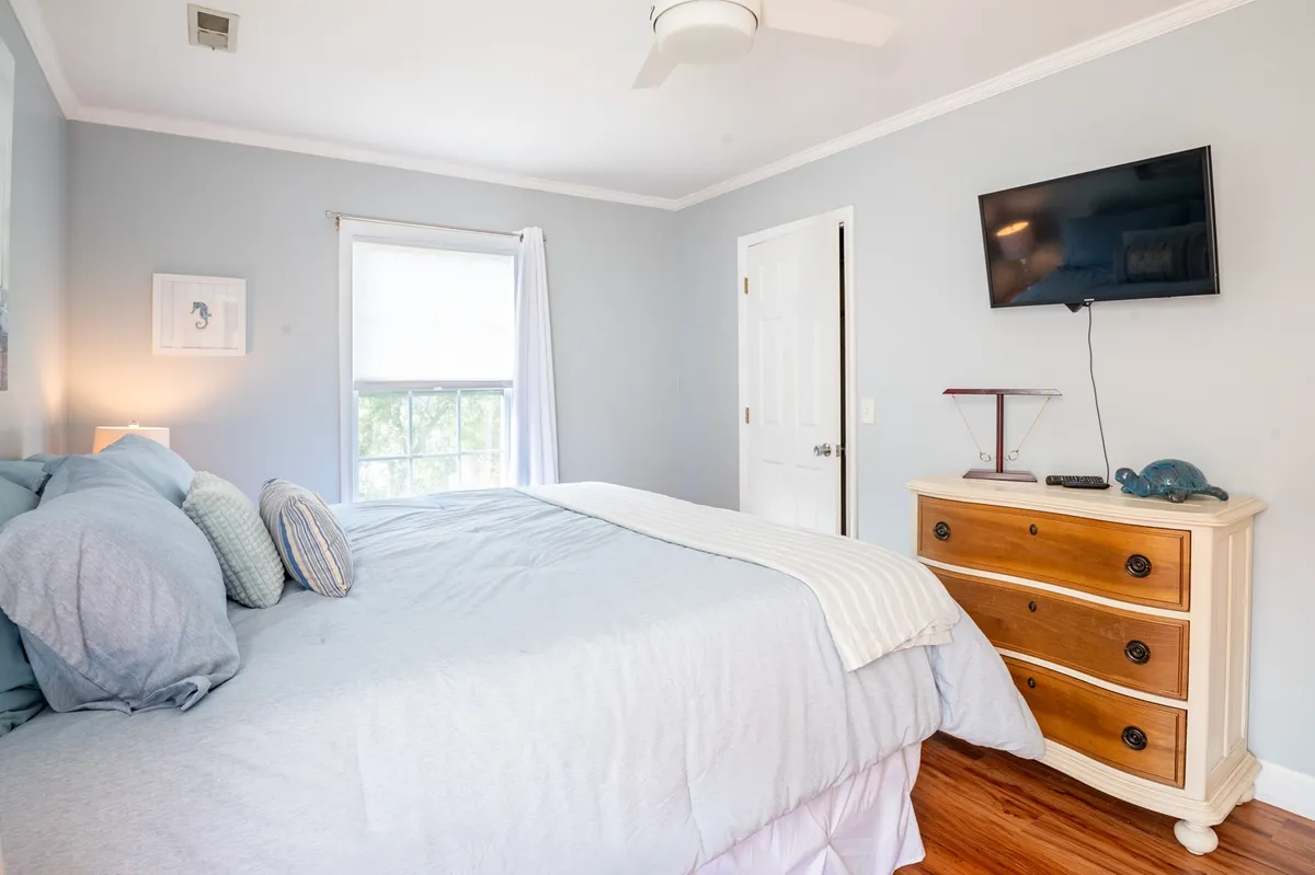 A comfortable guest bedroom featuring a bed with light blue linens, a wooden dresser, a wall-mounted TV, and a window that lets in gentle natural light. The room feels serene and welcoming—ideal for friends or family.