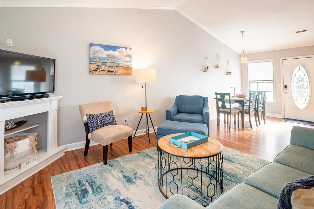 A cozy and welcoming great room featuring a plush sofa, coffee table, artwork, television, contemporary light fixture, dining table, and area rug. The open layout visually connects the living and dining spaces, allowing for seamless interaction and plenty of natural light.