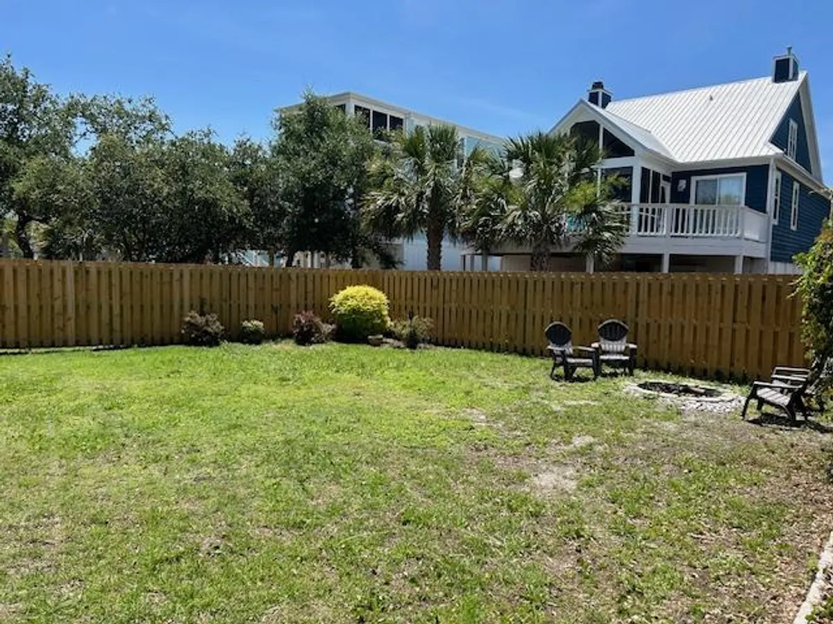 A lush backyard with a green lawn, wooden fence, palm trees, a cozy seating area with multiple chairs, and a small fire pit at the center. The environment feels inviting and is clearly designed for both play and communal gatherings.