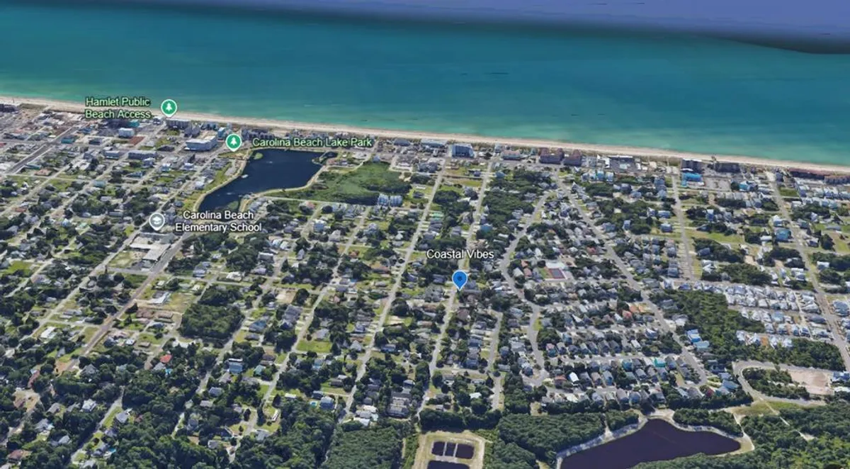 A sweeping view of Carolina Beach, with golden sands stretching out to meet the gentle waves of the Atlantic Ocean. In the foreground, the boardwalk hints at easy beach access, while blue skies and palm-lined paths convey an atmosphere perfect for family adventures, seashell hunts, and dolphin spotting.