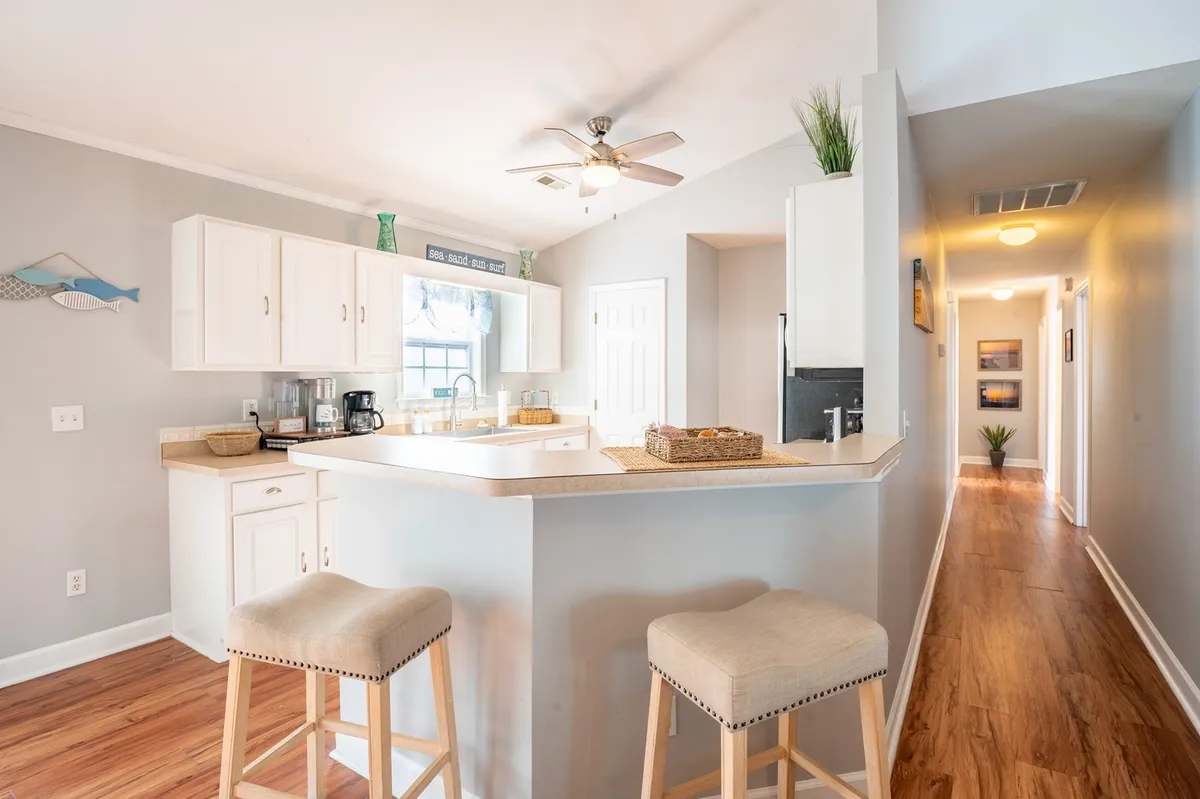 The image showcases a bright, inviting kitchen with a central island, stylish bar stools, and sunlight pouring in from a window. The open space encourages gathering, whether for morning coffee, preparing meals, or sharing conversation. Modern cabinetry and clean finishes set a relaxed coastal tone.