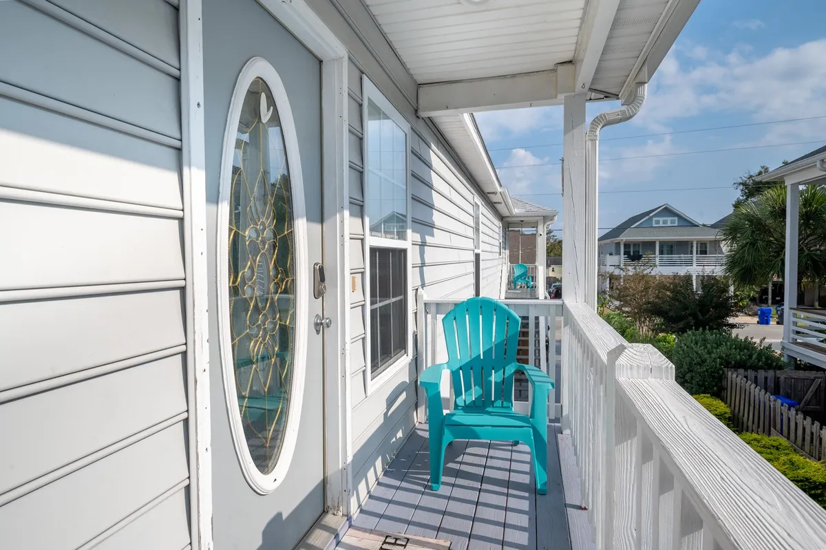 This exterior image features a bright turquoise chair placed beside a decorative front door, framed by a white railing. The space suggests a welcoming spot for morning coffee, relaxing, or greeting friends, with the outdoors visible and inviting.
